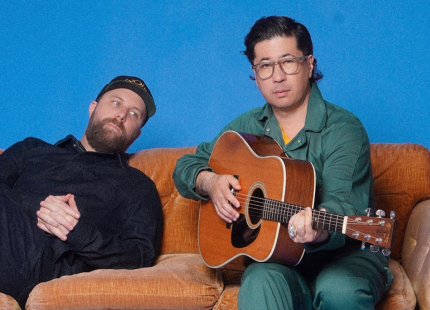 Daveed Goldman and Nobu Adilman pose on a sofa with a guitar.
