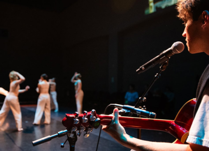 Guitarist plays on stage with dancers in the background.